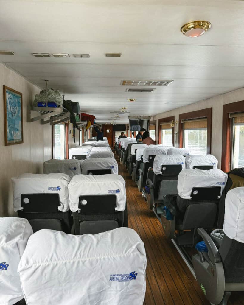 TABSA Ferry: Sailing from Puerto Natales to Puerto Yungay 12 Interior of the Tabsa Ferry in Chile showing a long aisle with cushioned seats covered in white linens labeled “Transbordadora Austral Broom,” with passengers stowing luggage above.