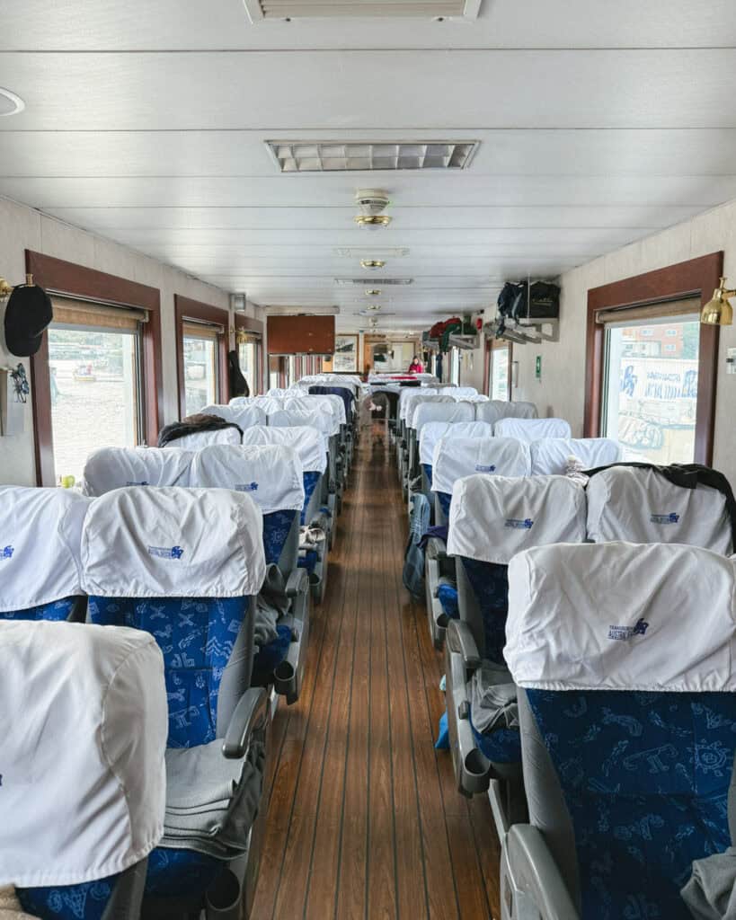 TABSA Ferry: Sailing from Puerto Natales to Puerto Yungay 23 Tabsa Ferry seating area in Chile with blue-patterned seats covered in branded white linens, wooden floors, and a central aisle leading toward the boarding area in the distance.