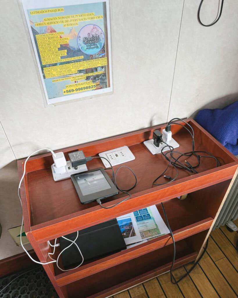 TABSA Ferry: Sailing from Puerto Natales to Puerto Yungay 33 Charging station aboard the Tabsa Ferry in Chile with multiple electronic devices plugged in, including a Kindle, surrounded by outlet strips on a wooden shelf below a poster advertising delivery service from Almacén Nómade in Puerto Edén.