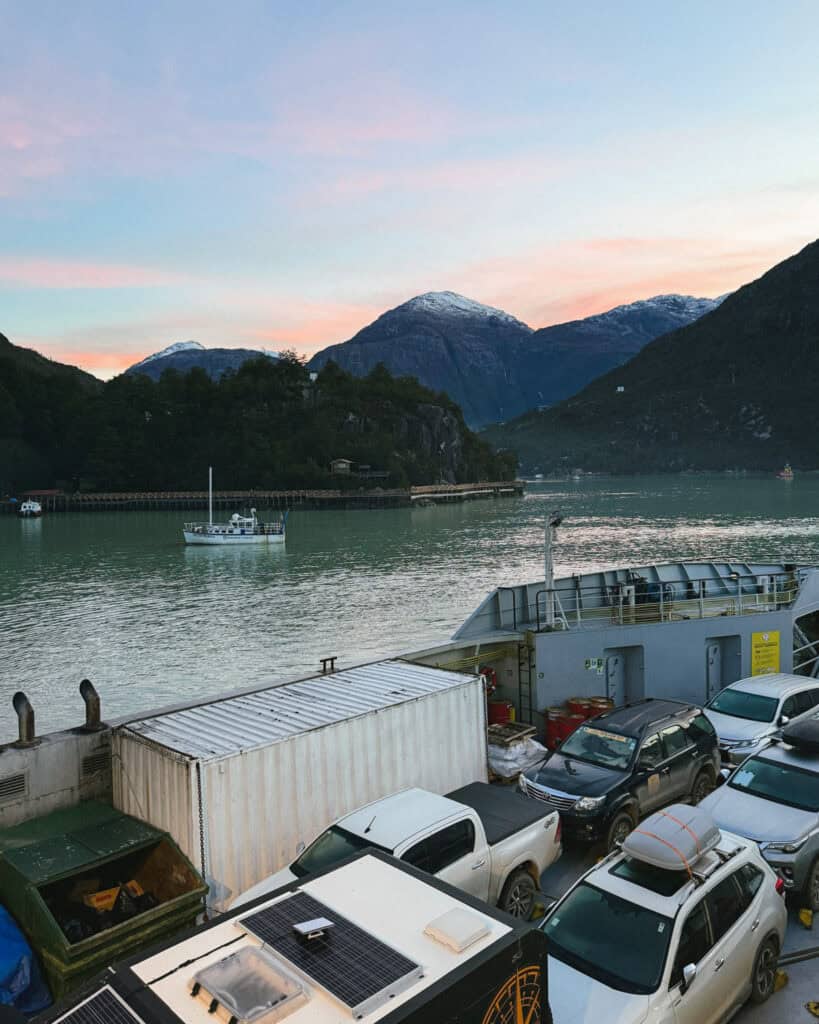TABSA Ferry: Sailing from Puerto Natales to Puerto Yungay 43 Scenic sunset view from the Tabsa Ferry in Chile, showing parked vehicles on the ferry deck and a snow-dusted mountain landscape reflected in the calm green water near Puerto Edén.