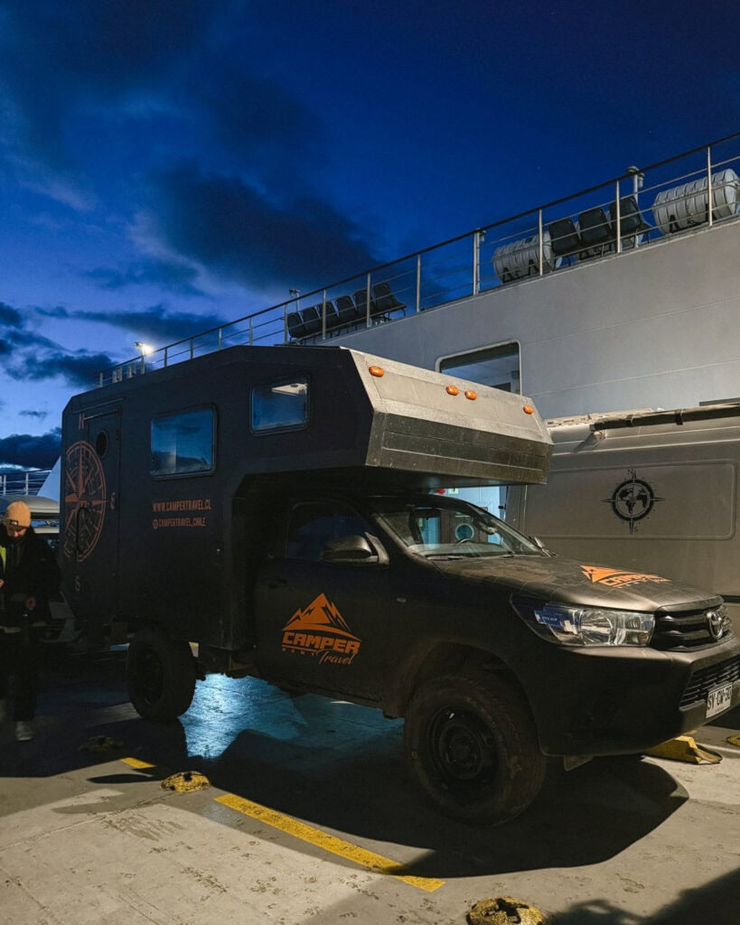 TABSA Ferry: Sailing from Puerto Natales to Puerto Yungay 22 Black Camper Travel vehicle parked on the deck of the Tabsa Ferry in Chile at night, with overhead lights casting shadows and a deep blue sky in the background.