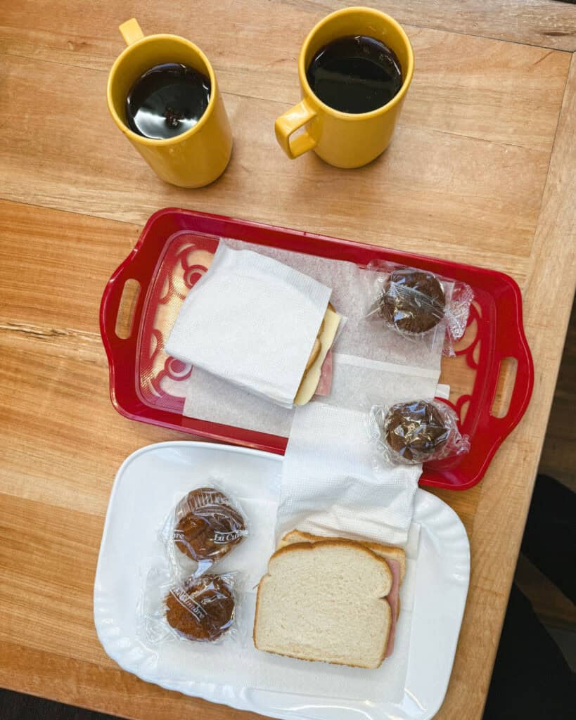 TABSA Ferry: Sailing from Puerto Natales to Puerto Yungay 13 Simple breakfast setup aboard the Tabsa Ferry in Chile, featuring two yellow mugs of coffee, two sandwiches wrapped in napkins, and packaged chocolate muffins on red and white trays placed on a wooden table.