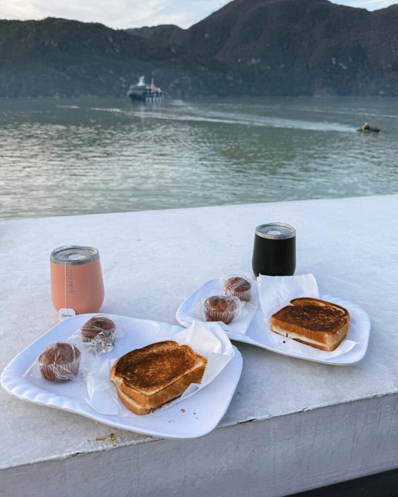 TABSA Ferry: Sailing from Puerto Natales to Puerto Yungay 29 Toasted breakfast sandwiches and packaged muffins served on white trays outdoors on the Tabsa Ferry in Chile, with tumblers of hot drinks and a scenic fjord view with distant ferry in the background.