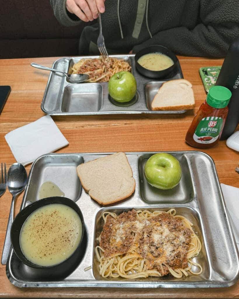 TABSA Ferry: Sailing from Puerto Natales to Puerto Yungay 27 Two metal trays with spaghetti bolognese, green apples, white bread slices, and creamy soup served aboard the Tabsa Ferry in Chile, with Brayden lifting his fork at the wooden table.