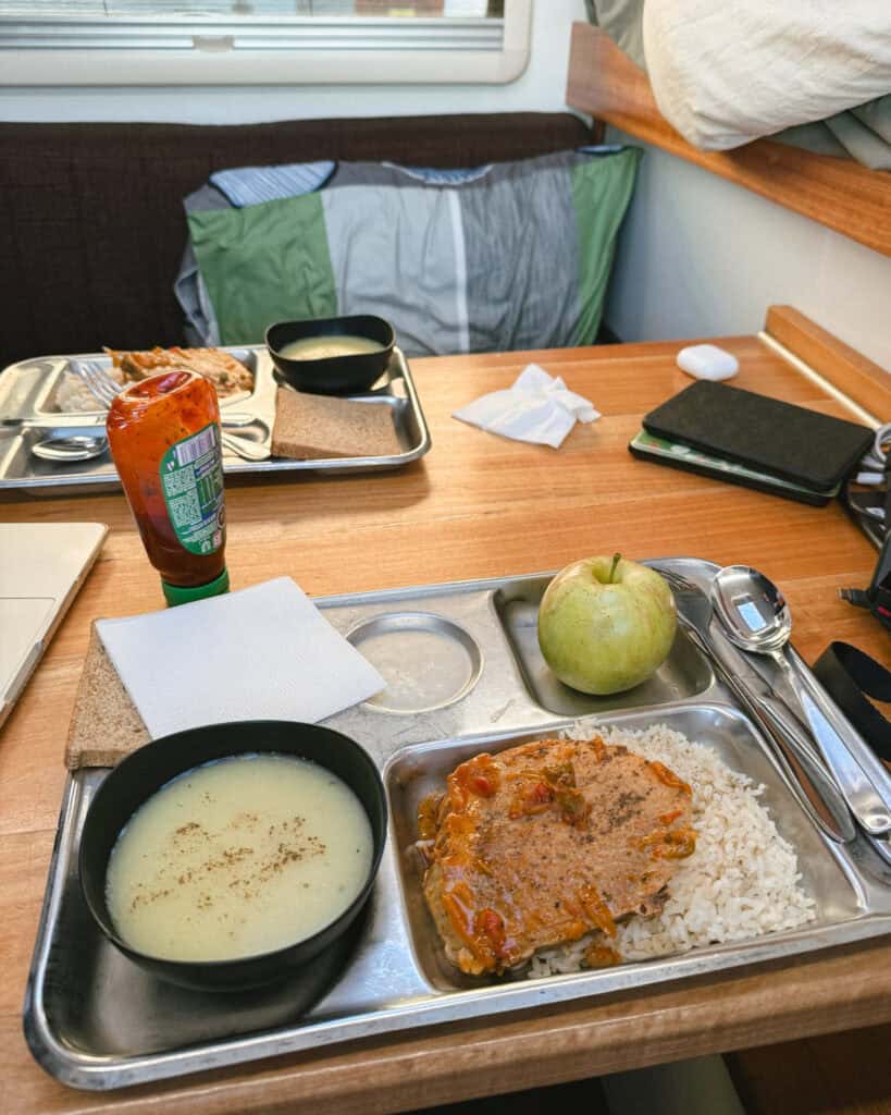 TABSA Ferry: Sailing from Puerto Natales to Puerto Yungay 30 Rice and vegetable patty served with creamy soup, white bread, and a green apple on a metal tray aboard the Tabsa Ferry in Chile, with a hot sauce bottle placed nearby on the wooden table.