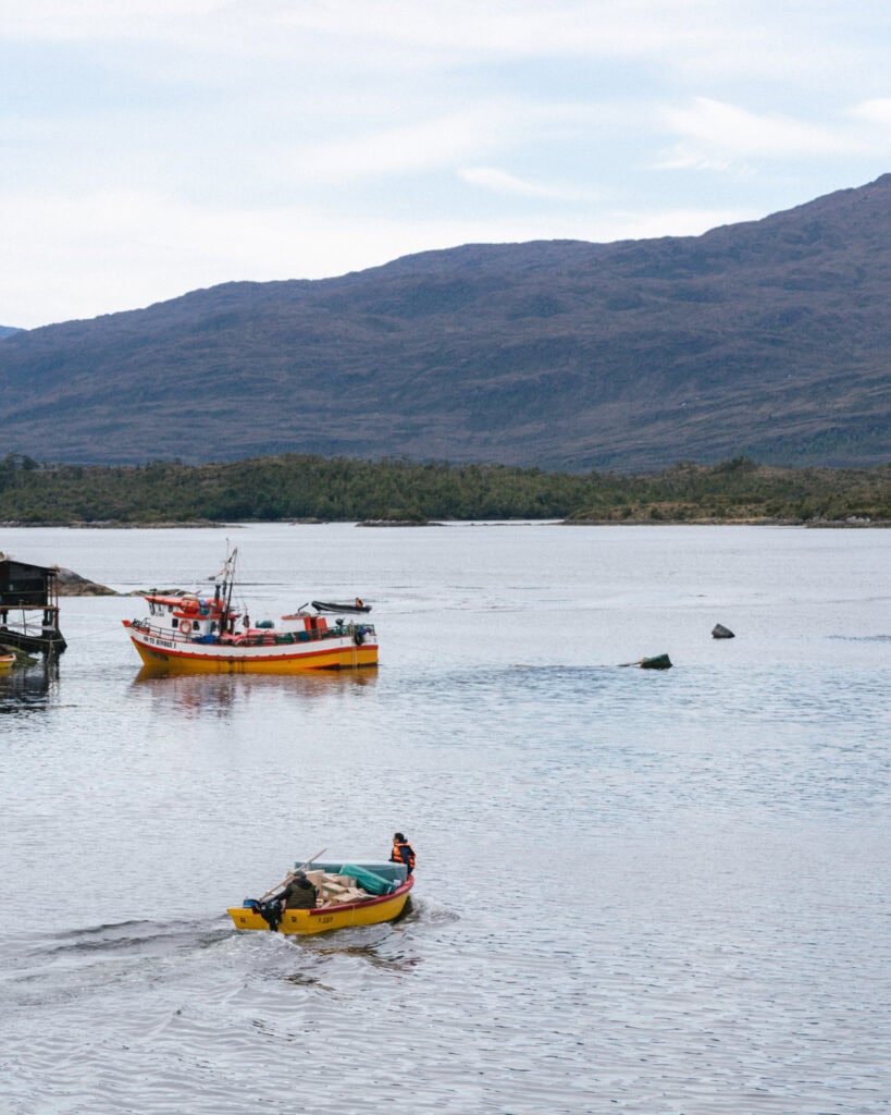 TABSA Ferry: Sailing from Puerto Natales to Puerto Yungay 44 Bright yellow and red fishing boats float in a calm bay near the Tabsa Ferry in Chile, with one small boat in the foreground carrying cargo and a person in an orange life jacket steering.