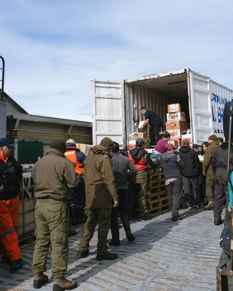 TABSA Ferry: Sailing from Puerto Natales to Puerto Yungay 45 Wide view of a group of workers and passengers, including people in military and safety uniforms, unloading stacked goods and produce boxes from a white cargo container at the Tabsa Ferry dock in Chile.
