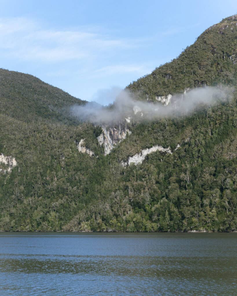 TABSA Ferry: Sailing from Puerto Natales to Puerto Yungay 38 Misty cloud drifting along a green mountainside covered in dense forest, with rock outcroppings and bright blue skies above, seen from the deck of the Tabsa Ferry.