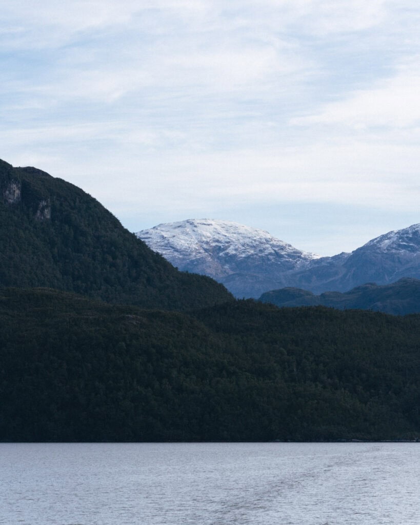 TABSA Ferry: Sailing from Puerto Natales to Puerto Yungay 7 Scenic view of forested hills with snow-capped mountains in the distance under a pale blue sky, captured from the Tabsa Ferry along Chile’s southern fjords.
