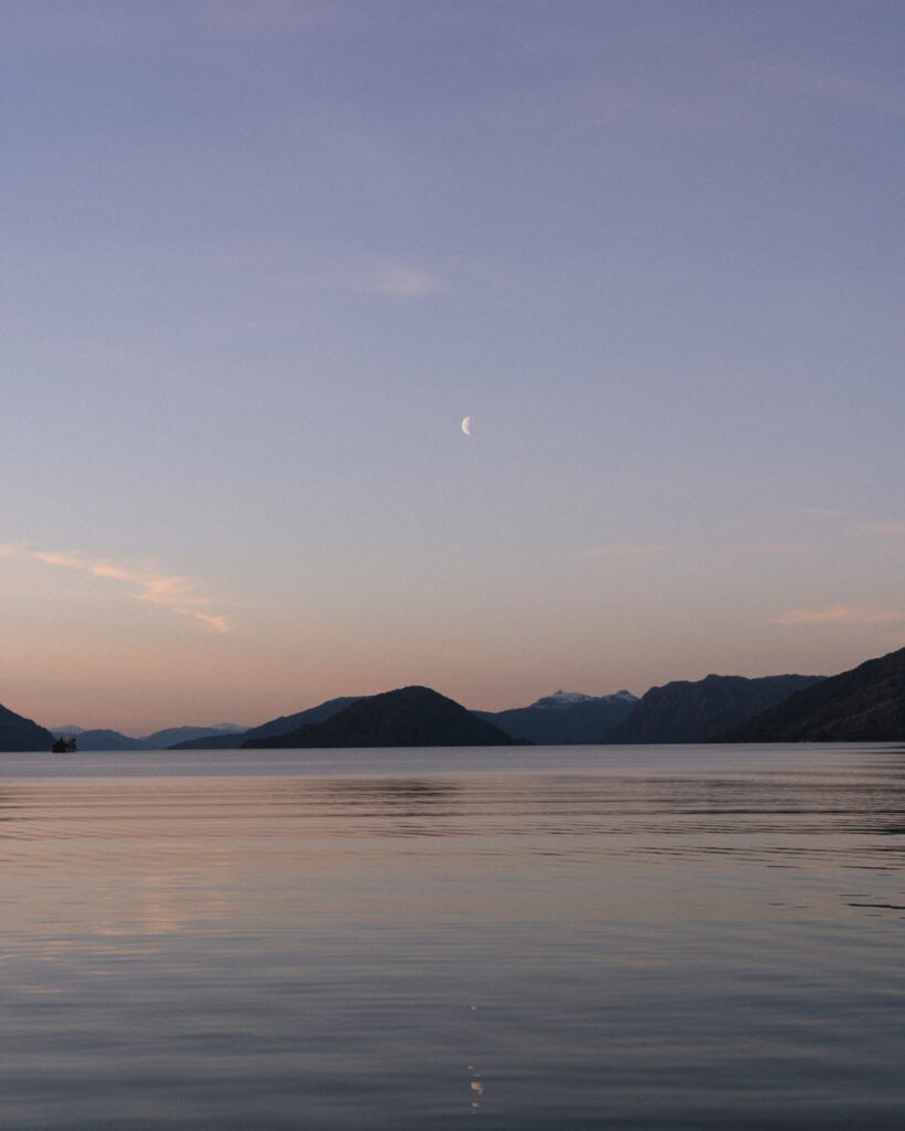 TABSA Ferry: Sailing from Puerto Natales to Puerto Yungay 41 Soft pastel twilight over the fjords of southern Chile, with a crescent moon hanging above the silhouette of mountains and calm reflective waters during sunset.