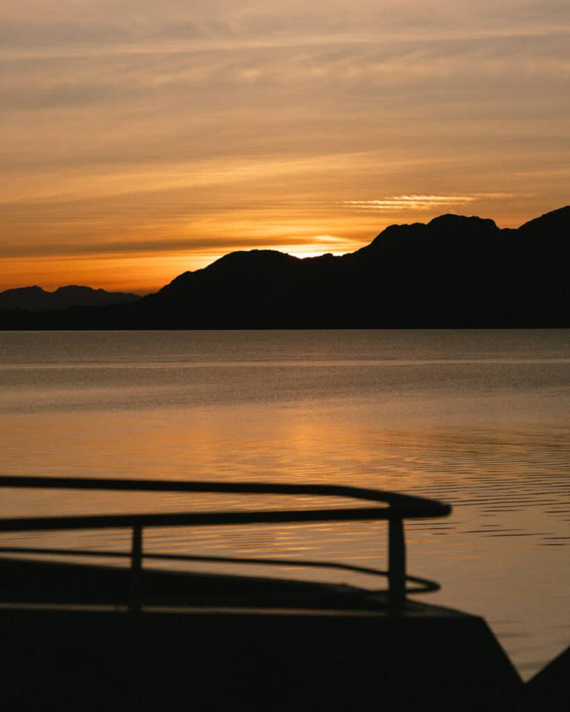 TABSA Ferry: Sailing from Puerto Natales to Puerto Yungay 37 Golden-orange hues of sunset over tranquil waters and dark silhouetted mountains, with a blurred ferry railing in the foreground.