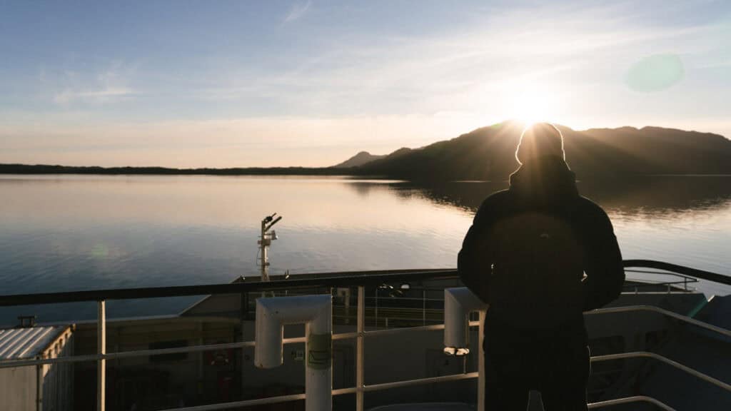 TABSA Ferry: Sailing from Puerto Natales to Puerto Yungay 46 Brayden in a hooded jacket silhouetted against the rising sun, standing at the railing of the Tabsa Ferry, overlooking still waters and distant hills at dawn.