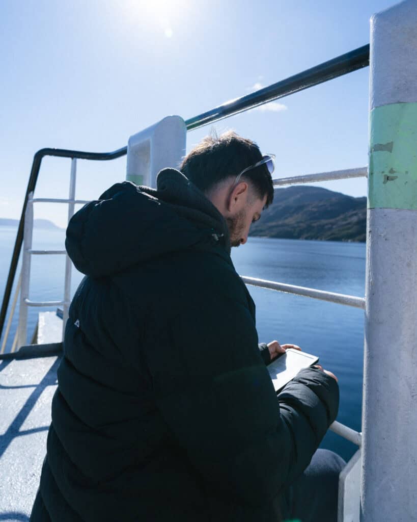 TABSA Ferry: Sailing from Puerto Natales to Puerto Yungay 36 Brayden sits on the outer deck of the Tabsa Ferry reading a book, wrapped in a black puffer jacket with the sun shining overhead and the fjord waters glistening behind him.