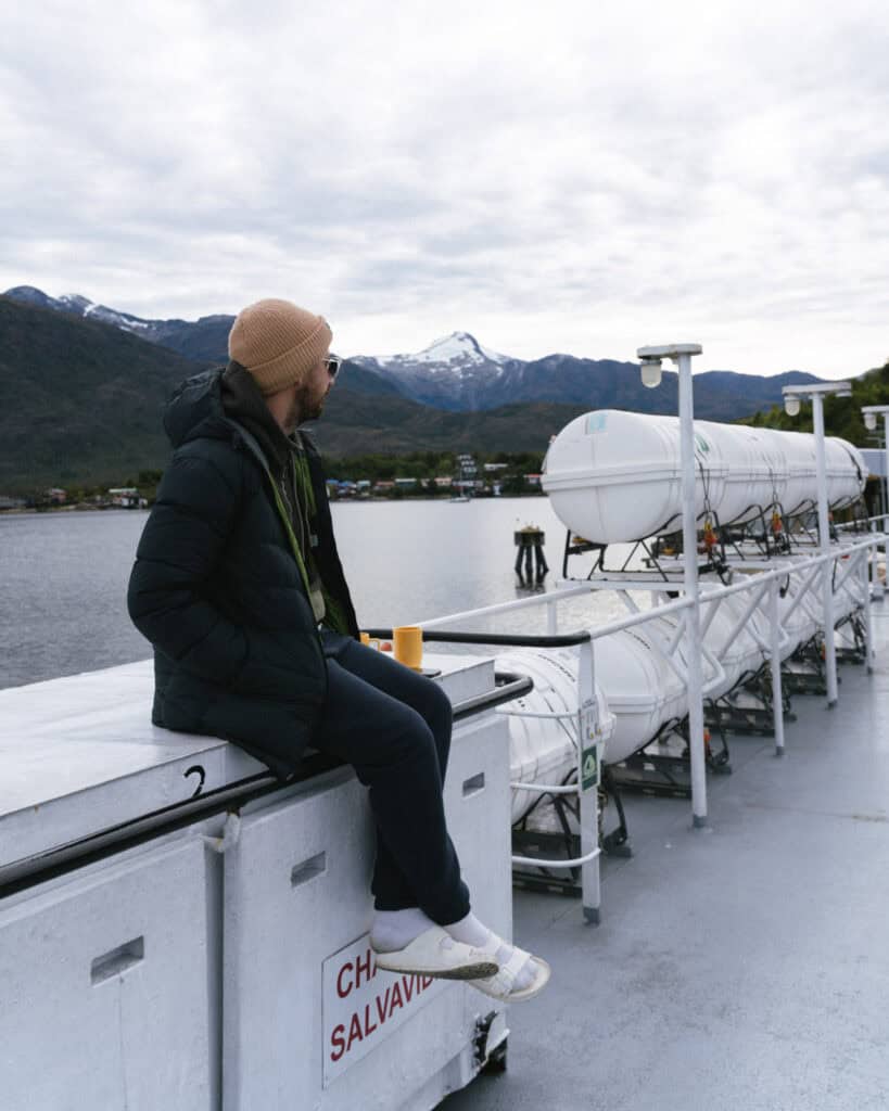 TABSA Ferry: Sailing from Puerto Natales to Puerto Yungay 20 Brayden in a tan beanie and dark coat sits on the ferry deck railing with a coffee mug beside him, overlooking a mountain village surrounded by snowy peaks during a stop.