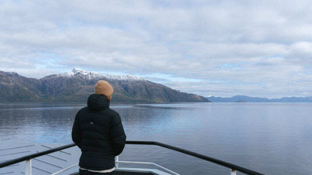 TABSA Ferry: Sailing from Puerto Natales to Puerto Yungay 15 Brayden stands at the ferry’s bow in a tan beanie and black coat, gazing toward dramatic snow-capped peaks and cloud-speckled skies along the fjord route from Puerto Natales.