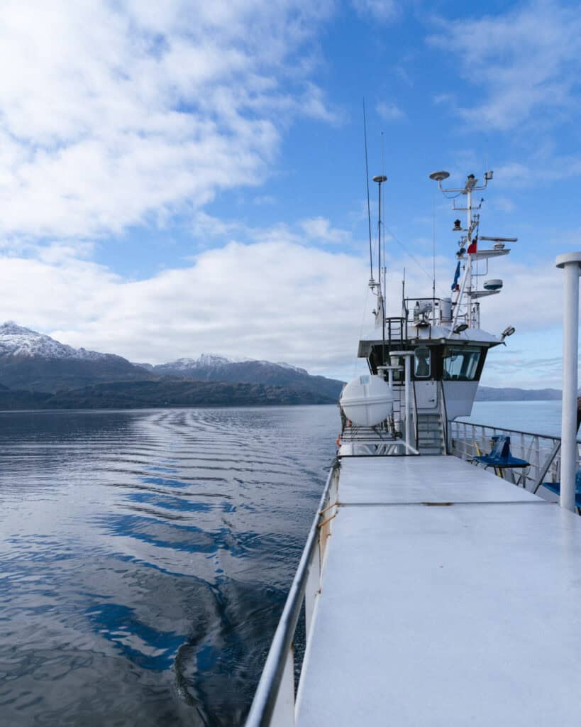 TABSA Ferry: Sailing from Puerto Natales to Puerto Yungay 2 View from the side deck of the ferry, showing rippling wake on the water, the ship’s command tower, and snow-capped mountains under partly cloudy skies.