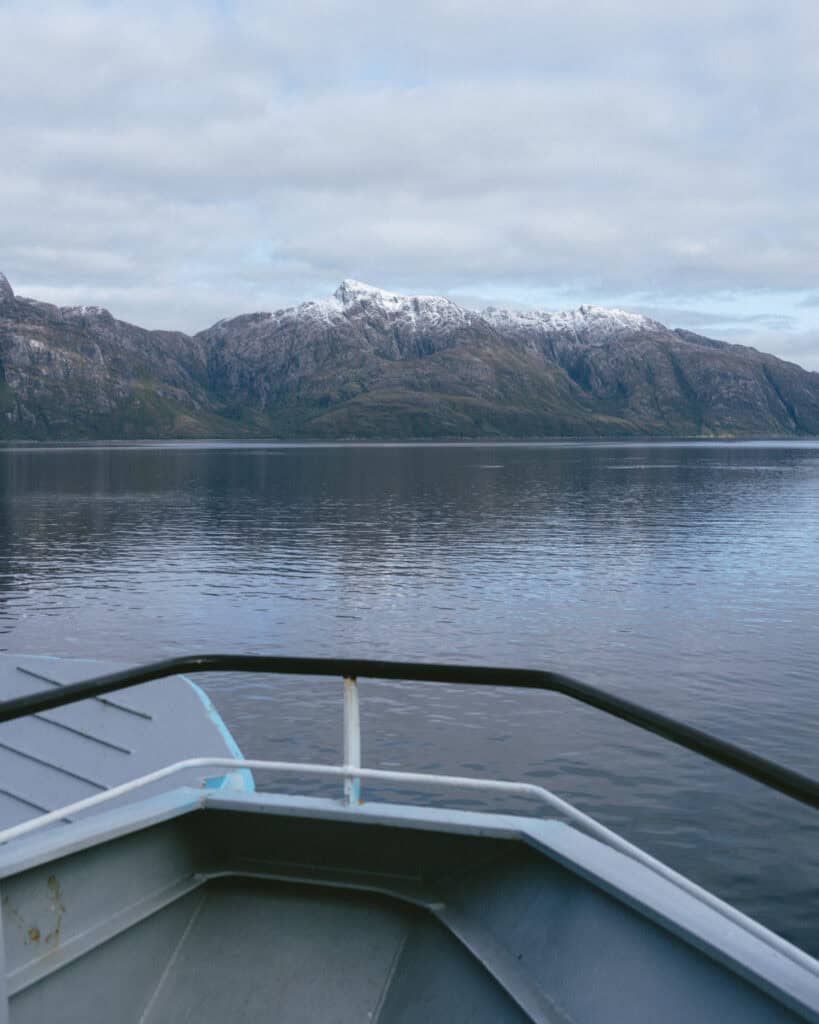 TABSA Ferry: Sailing from Puerto Natales to Puerto Yungay 39 View over the front of the ferry, showing peaceful waters and towering rocky mountains capped with snow, beneath a moody, overcast sky.