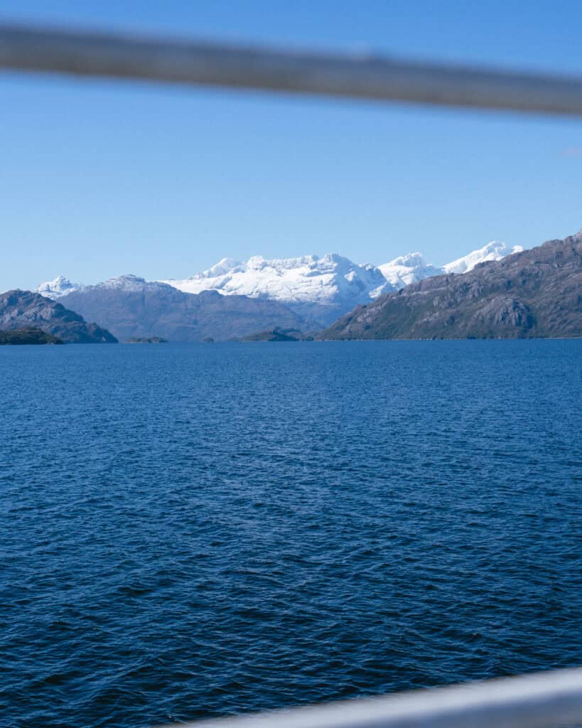 TABSA Ferry: Sailing from Puerto Natales to Puerto Yungay 40 Snow-draped Patagonian peaks captured through a partially obscured view framed by the ferry's metal railing in the foreground, under clear blue skies.