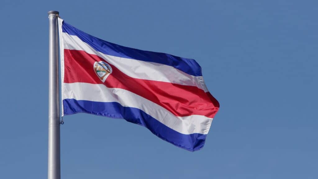 Costa Rica Travel Guide: Planning The Perfect Pura Vida Trip 32 An image of the Costa Rica flag on a flagpole flying against a blue sky. The flag has two horizontal stripes on the top and bottom, followed by two white stripes, meeting at a single red stripe along the centre with a small logo on the left side.