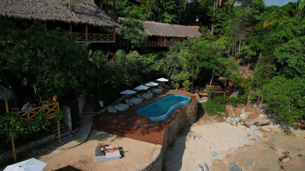 Quimixto Travel Guide: Puerto Vallarta's Hidden Gem 18 Secluded beachfront resort in Quimixto, Mexico, featuring a small elevated pool with lounge chairs and umbrellas nestled among lush jungle foliage. Thatched-roof palapas and rustic cabins are surrounded by dense greenery, with a sandy beach and stone pathway leading to the ocean.
