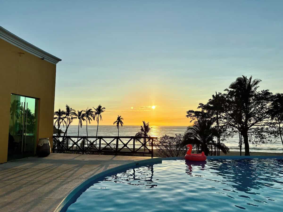 San Pancho Mexico Travel Guide: Tropical Bliss in Nayarit 11 Infinity pool at sunset overlooking the Pacific Ocean in San Pancho, Nayarit, Mexico, with a pink flamingo pool float gently drifting on the water. Silhouettes of palm trees frame the golden sky, while the ocean reflects the setting sun behind a wooden fence along the beachfront.