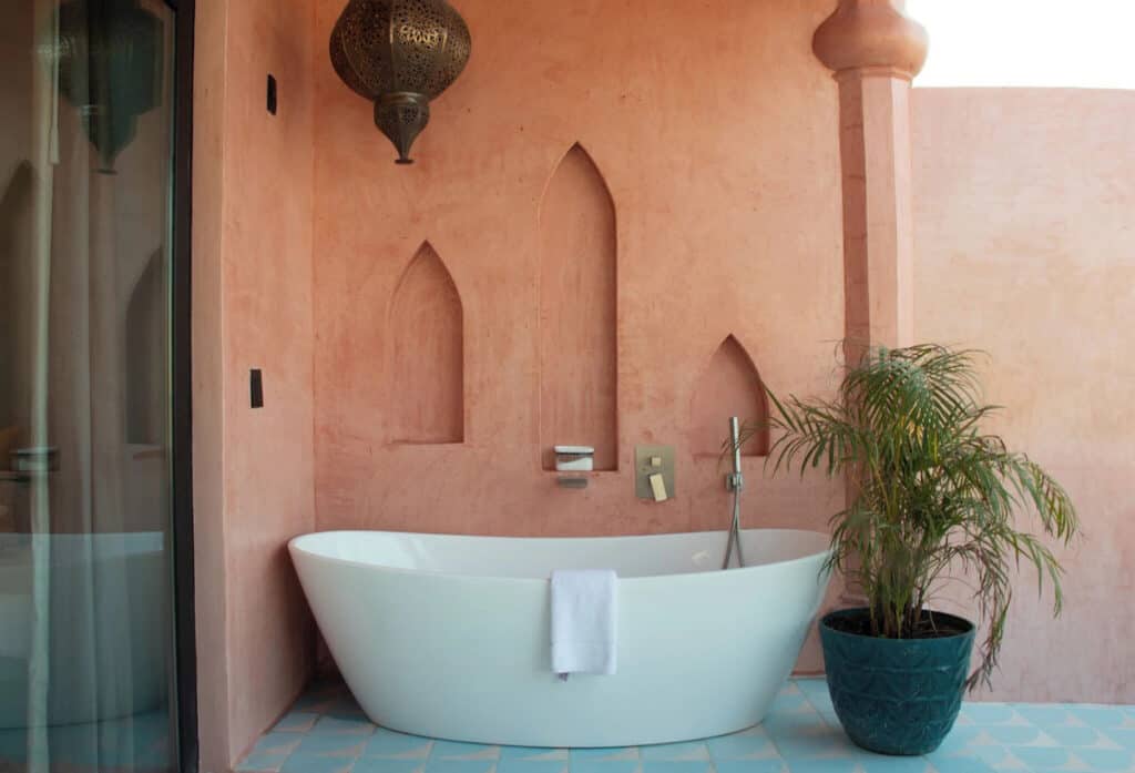 Sayulita Travel Guide: Nayarit, Mexico's Buzzing Surf Town 10 Freestanding white bathtub set against a terracotta-pink stucco wall with arched alcoves and a decorative hanging lantern in a Moroccan-inspired outdoor bathroom in Nomada Wellness Hotel in Sayulita, Mexico. The space features light blue tile flooring, a potted palm plant, and brass fixtures, blending bohemian and spa-like design elements.