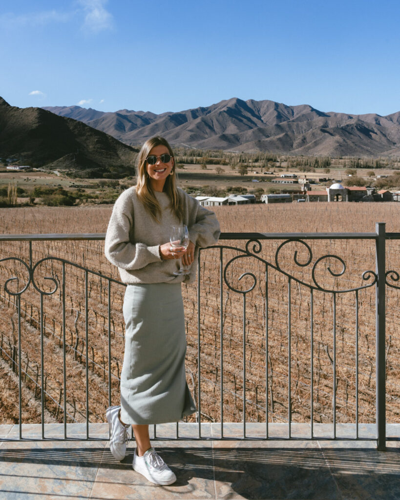7 - 10 Day Salta Road Trip: The Best of Northwest Argentina 46 Sally stands smiling with a wine glass in hand on a scenic balcony in Cachi, surrounded by a vineyard and backed by dramatic mountain views.