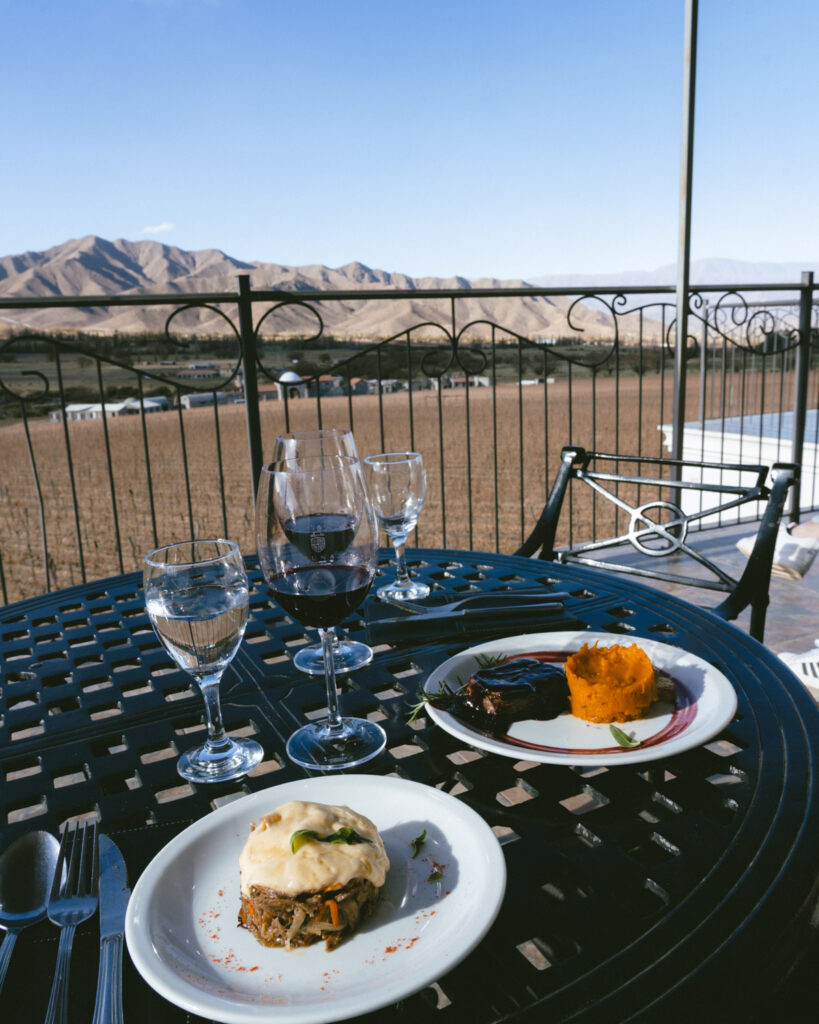 7 - 10 Day Salta Road Trip: The Best of Northwest Argentina 57 A vineyard-facing lunch table displays two elegant main courses—a vegetable tower topped with cheese and a steak with mashed sweet potatoes—paired with red wine and water.