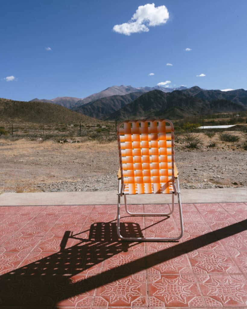 7 - 10 Day Salta Road Trip: The Best of Northwest Argentina 60 An orange woven lawn chair casts a shadow on a red-tiled porch floor, facing a mountain range under a bright blue sky.