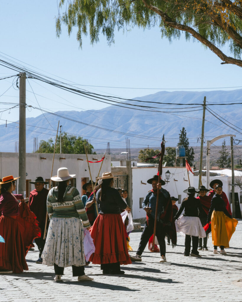 7 - 10 Day Salta Road Trip: The Best of Northwest Argentina 50 A group of traditionally dressed dancers in colorful skirts and wide-brimmed hats perform a folk dance on a cobbled street in Cachi, with the Andes mountains behind them.