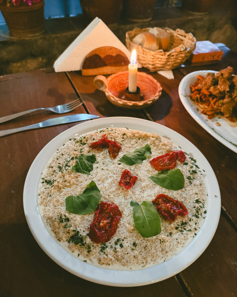 7 - 10 Day Salta Road Trip: The Best of Northwest Argentina 55 A creamy quinoa dish garnished with sun-dried tomatoes and fresh green leaves is served on a white plate beside a lit candle and bread basket.