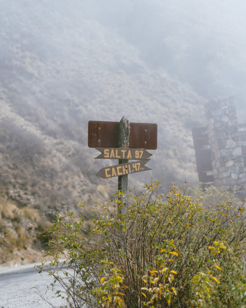7 - 10 Day Salta Road Trip: The Best of Northwest Argentina 67 A wooden road sign in the fog marks distances to Salta (97 km) and Cachi (47 km), standing beside a bush on a misty roadside.