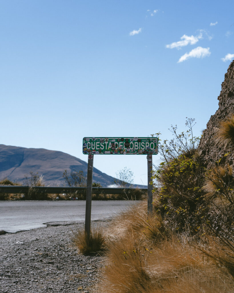 7 - 10 Day Salta Road Trip: The Best of Northwest Argentina 65 A green road sign reading “Cuesta del Obispo” is covered in stickers and stands beside a winding mountain road under a clear sky.