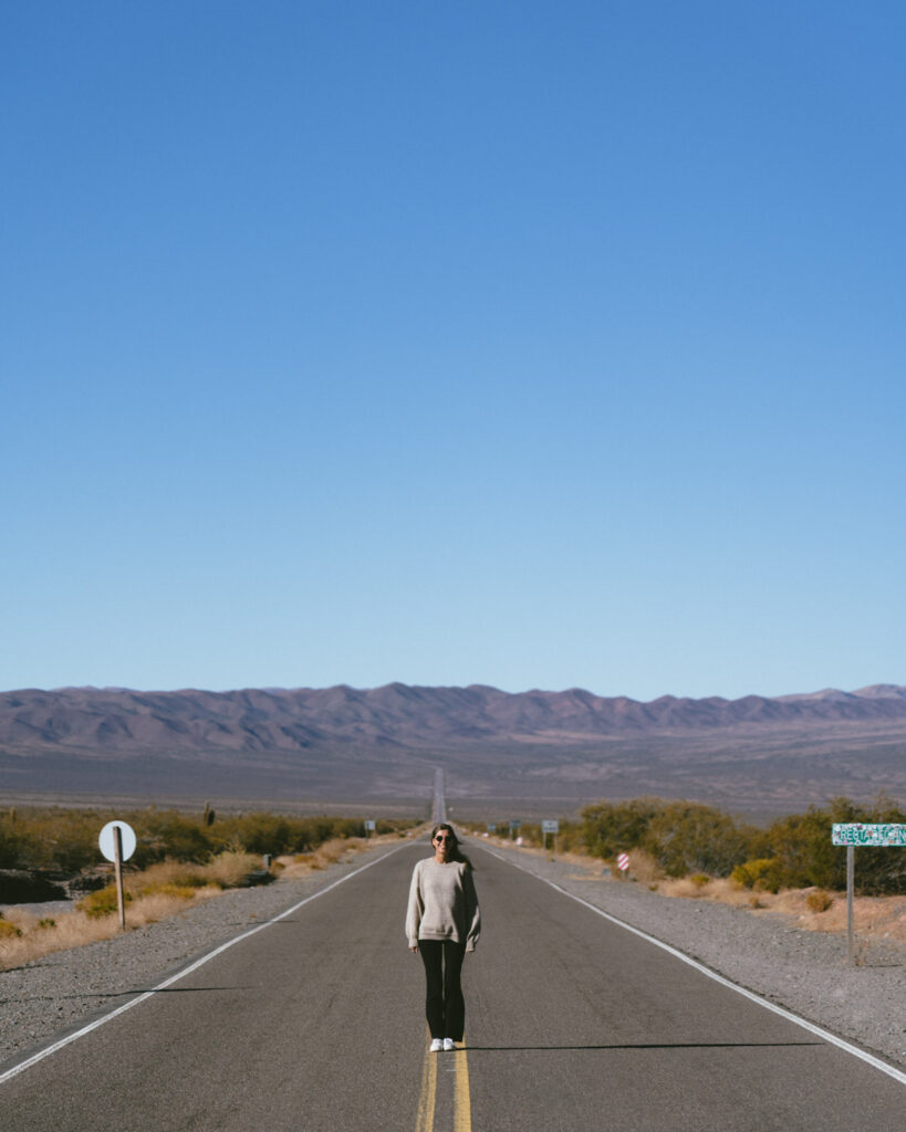 7 - 10 Day Salta Road Trip: The Best of Northwest Argentina 63 Sally in a gray sweater stands on the yellow center line of an empty road with desert landscape and mountains stretching behind her.