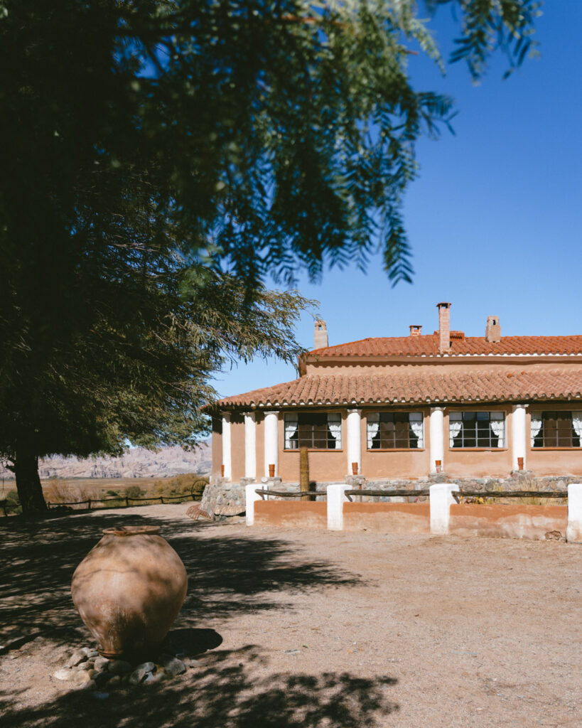 7 - 10 Day Salta Road Trip: The Best of Northwest Argentina 42 Clay-colored building with red tile roof partially framed by tree branches, with a large ceramic pot sitting on the sandy ground in the foreground.