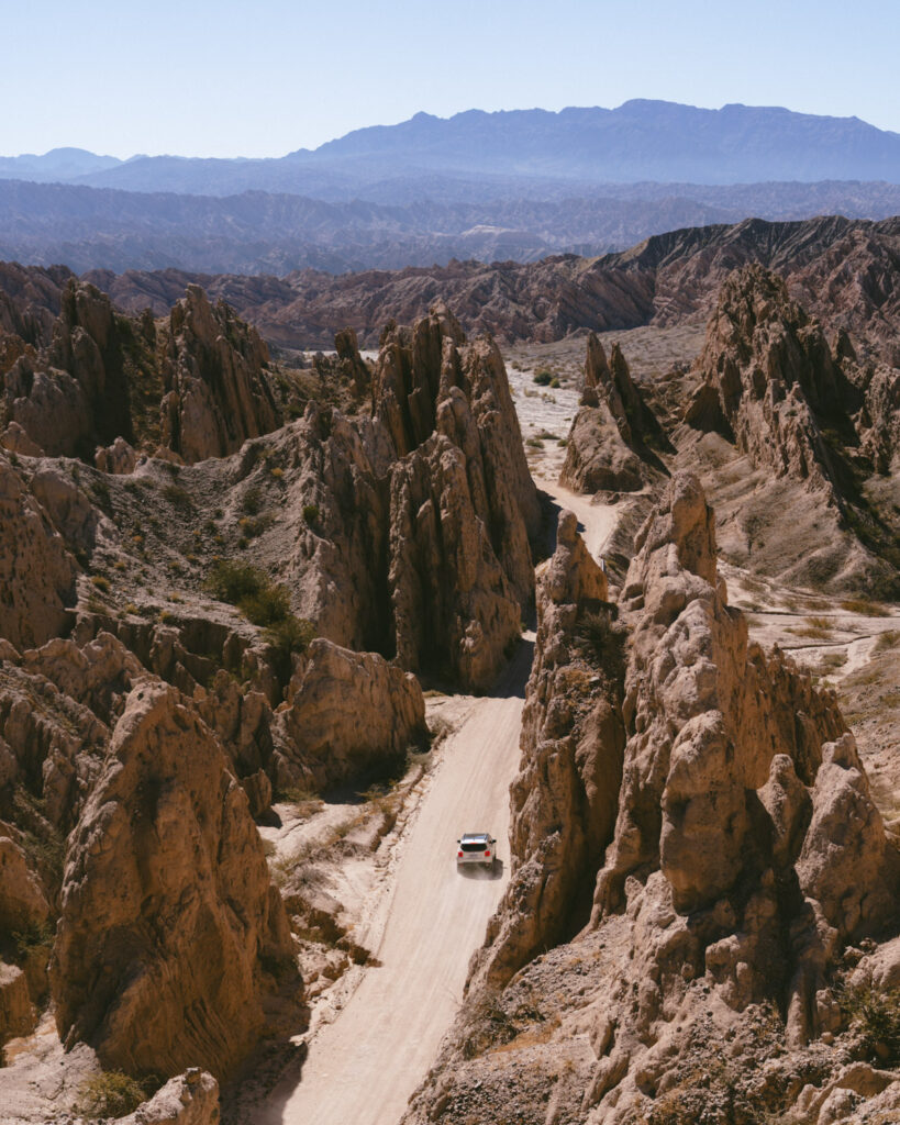 7 - 10 Day Salta Road Trip: The Best of Northwest Argentina 2 A small white car leaves a rugged rock passage, trailing dust along a desert road flanked by jagged cliffs and expansive mountainous terrain.
