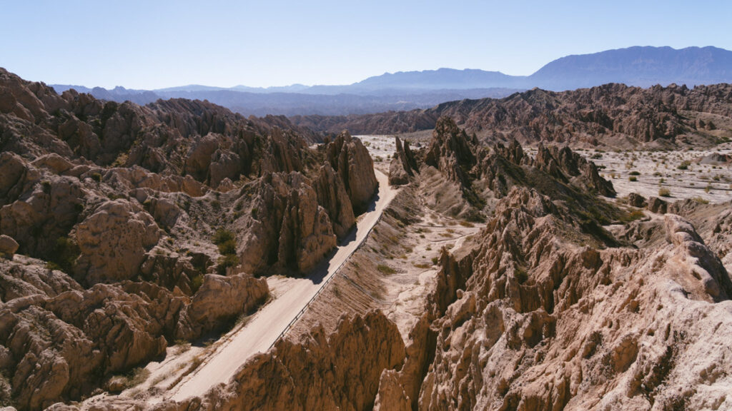 7 - 10 Day Salta Road Trip: The Best of Northwest Argentina 44 Panoramic view of a dirt road cutting through jagged desert rock formations with mountains fading into the horizon under a clear blue sky.