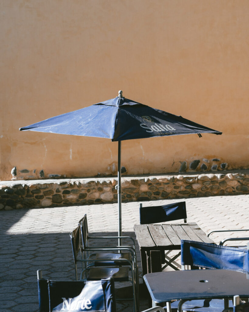 7 - 10 Day Salta Road Trip: The Best of Northwest Argentina 53 Outdoor café patio with a navy blue umbrella that reads “Salta” surrounded by empty metal-framed chairs and rustic stone walls.