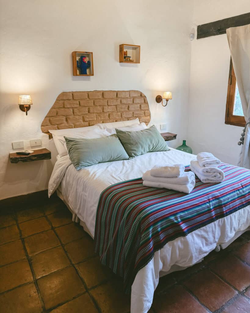 7 - 10 Day Salta Road Trip: The Best of Northwest Argentina 39 A cozy bedroom features a rustic bed with a woven Andean blanket, folded white towels, and soft lighting against white stucco walls.