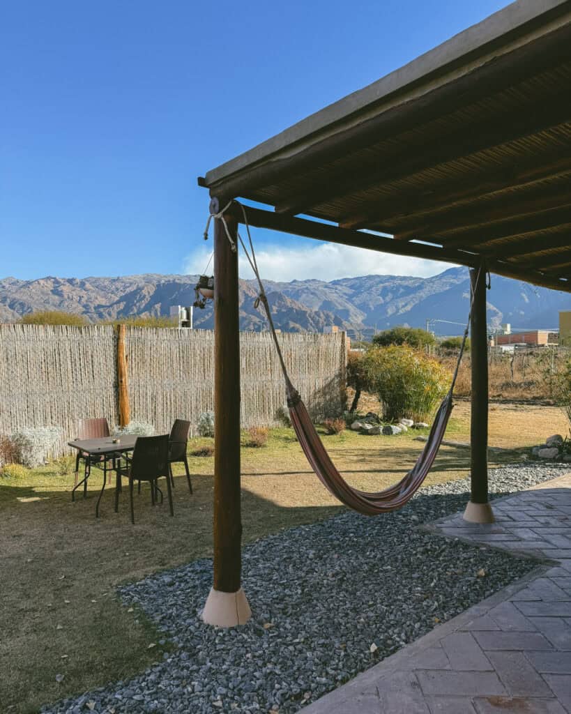 7 - 10 Day Salta Road Trip: The Best of Northwest Argentina 40 A striped hammock hangs under a shaded wooden patio, overlooking a backyard with a table and chairs, and the Andes mountains in the background.