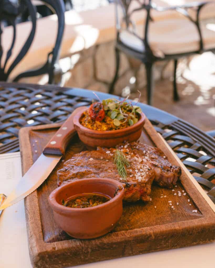 7 - 10 Day Salta Road Trip: The Best of Northwest Argentina 32 A seared steak topped with salt crystals is paired with two clay dishes of chimichurri and squash risotto on a rustic wood platter at an outdoor table.