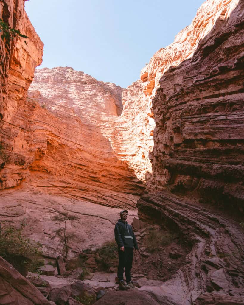 7 - 10 Day Salta Road Trip: The Best of Northwest Argentina 3 Brayden wearing a dark jacket and cap stands on rocky terrain inside a steep canyon with layered red cliffs rising high behind him.
