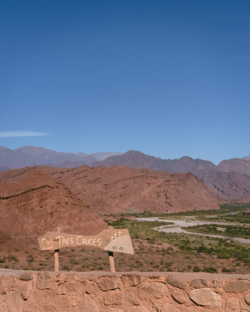 7 - 10 Day Salta Road Trip: The Best of Northwest Argentina 22 A frontal view of the “Tres Cruces” viewpoint sign with painted symbols for a camera and crosses, overlooking a vast, layered desert landscape.
