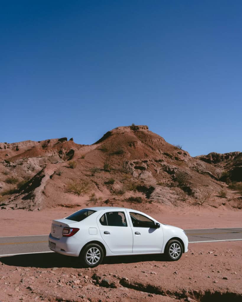 7 - 10 Day Salta Road Trip: The Best of Northwest Argentina 5 A white sedan is parked beside a road in a dry, rocky desert landscape with red hills under a bright blue sky.