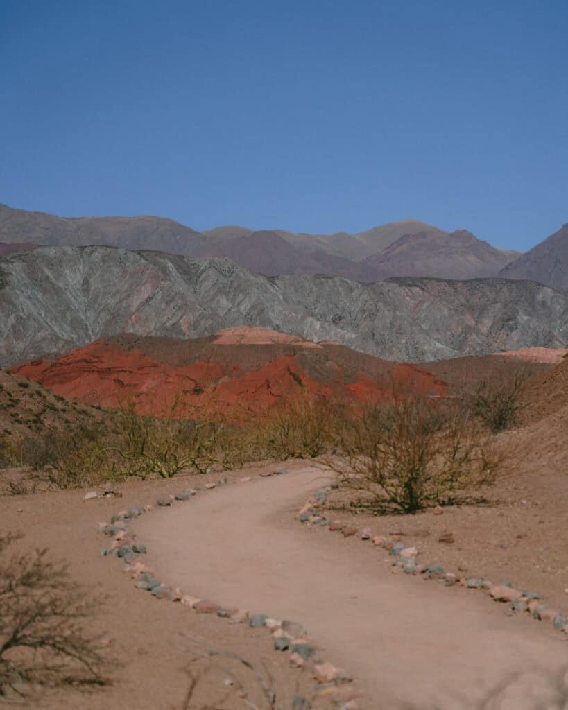 7 - 10 Day Salta Road Trip: The Best of Northwest Argentina 21 A sandy path curves gently through desert terrain with strikingly striped, multicolored mountains rising in the background.