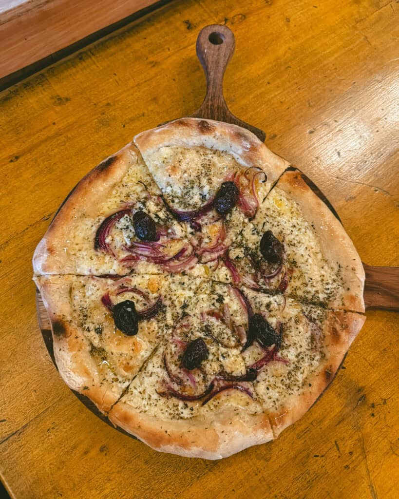 7 - 10 Day Salta Road Trip: The Best of Northwest Argentina 35 A fresh pizza topped with red onions, olives, and herbs is served on a wooden paddle on a warm wooden table.