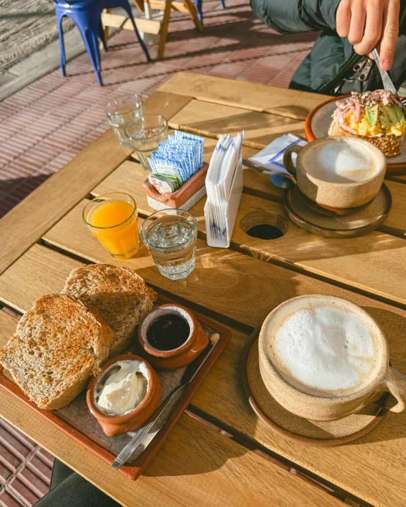 7 - 10 Day Salta Road Trip: The Best of Northwest Argentina 37 A wooden café table is set with cappuccinos, orange juice, toast with cream cheese and jam, and a colorful open-faced sandwich topped with avocado, ham, and seeds.