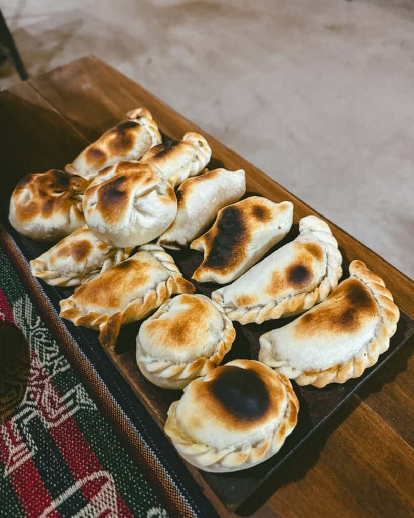 7 - 10 Day Salta Road Trip: The Best of Northwest Argentina 34 A wooden tray is filled with a dozen golden-browned empanadas on a woven tablecloth with traditional patterns.