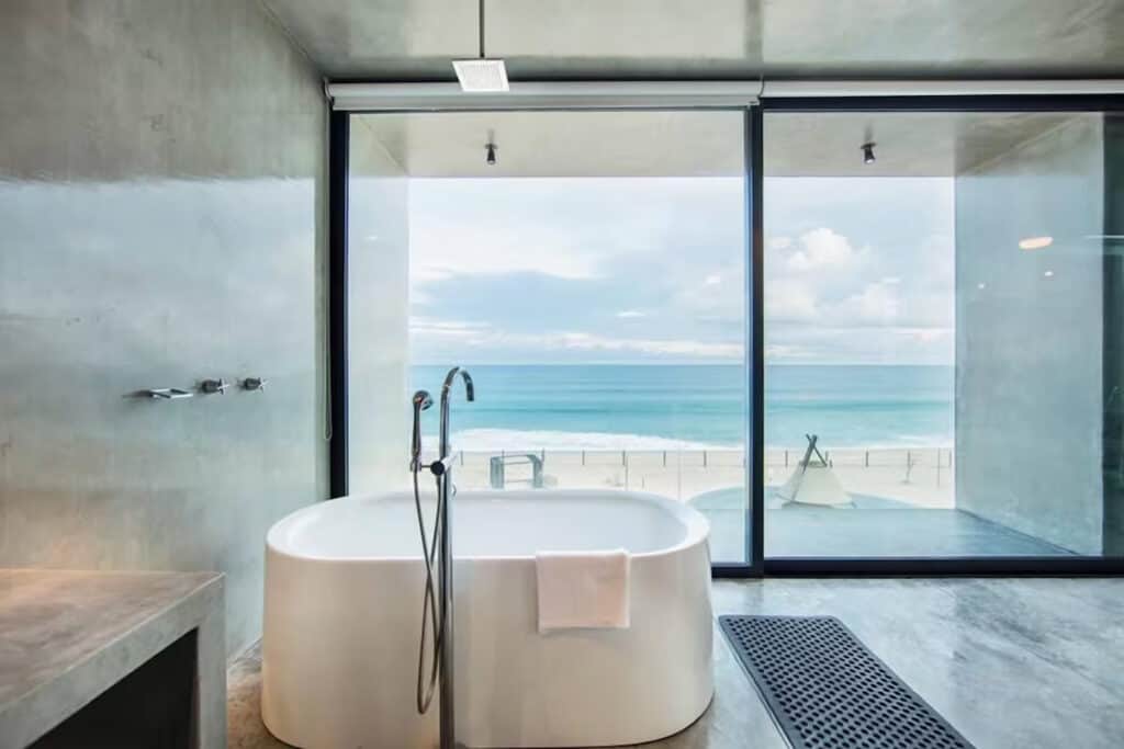East Cape Baja Travel Guide: Remote & Unspoiled Paradise 28 Modern, minimalist bathroom with a freestanding white bathtub facing floor-to-ceiling glass windows that overlook a serene beach and turquoise ocean in Vidasoul Hotel and Crossroads Country Club Restaurant Baja California Sur. The space features sleek concrete walls and floors with a luxurious, open-concept design.