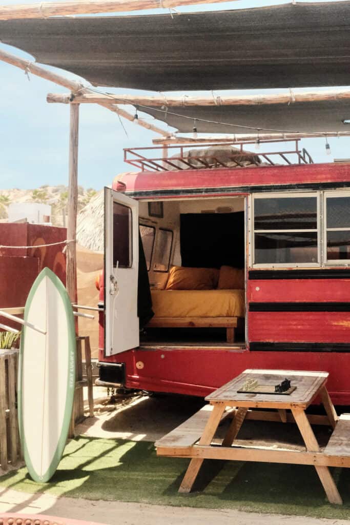 East Cape Baja Travel Guide: Remote & Unspoiled Paradise 27 The Baja Station, a unique East Cape accommodation featuring a converted red school bus outfitted with a cozy yellow-bedded interior. Outside, a wooden picnic table with a chessboard and a surfboard enhance the relaxed, beachside atmosphere of Baja California Sur.